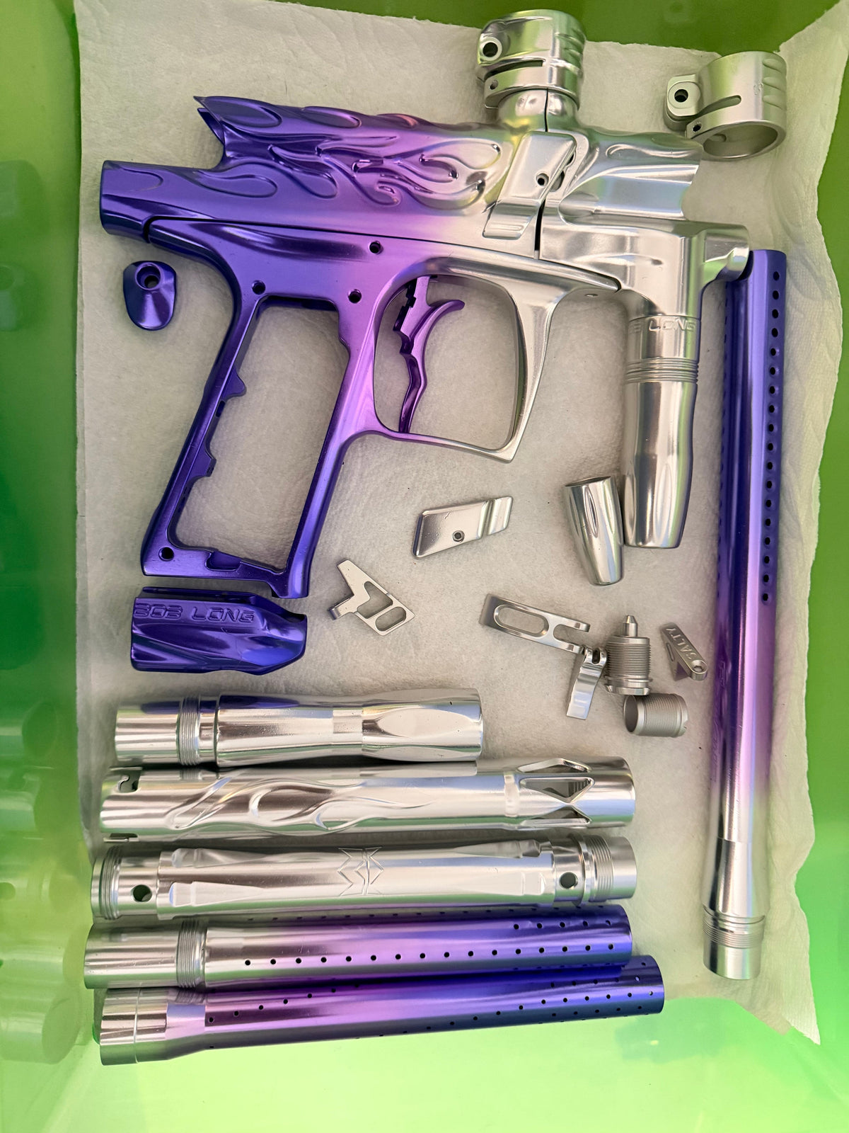 A complete paintball marker stripped into its parts, anodized in a purple-to-silver fade, laid out in a tray